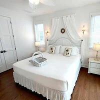 A bright bedroom with a white bed, two wall lamps, and two windows. A basket is placed on the bed. The room has wooden floors and a ceiling fan.