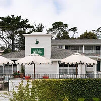 Exterior view of a two-story building with white walls, surrounded by green shrubs and trees, featuring patio umbrellas and a sign with text.