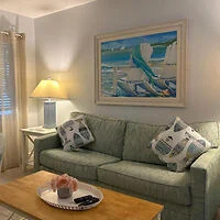 A cozy living room with a green sofa, two patterned pillows, a wooden coffee table with a tray, and a lamp. A painting depicting an ocean scene hangs on the wall.