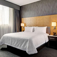 A hotel room with a neatly made bed, white linens, wooden headboard, bedside tables with lamps, and a window with curtains.