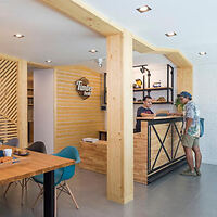 Two people converse at a wooden reception desk in a modern, minimalist cafe with light wood decor and contemporary furniture.