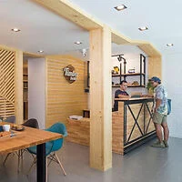 Two people converse at a wooden reception desk in a modern, minimalist cafe with light wood decor and contemporary furniture.