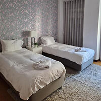 Two neatly made single beds with white linens in a room with a patterned accent wall, a gray carpet, and a small nightstand with a lamp between the beds.