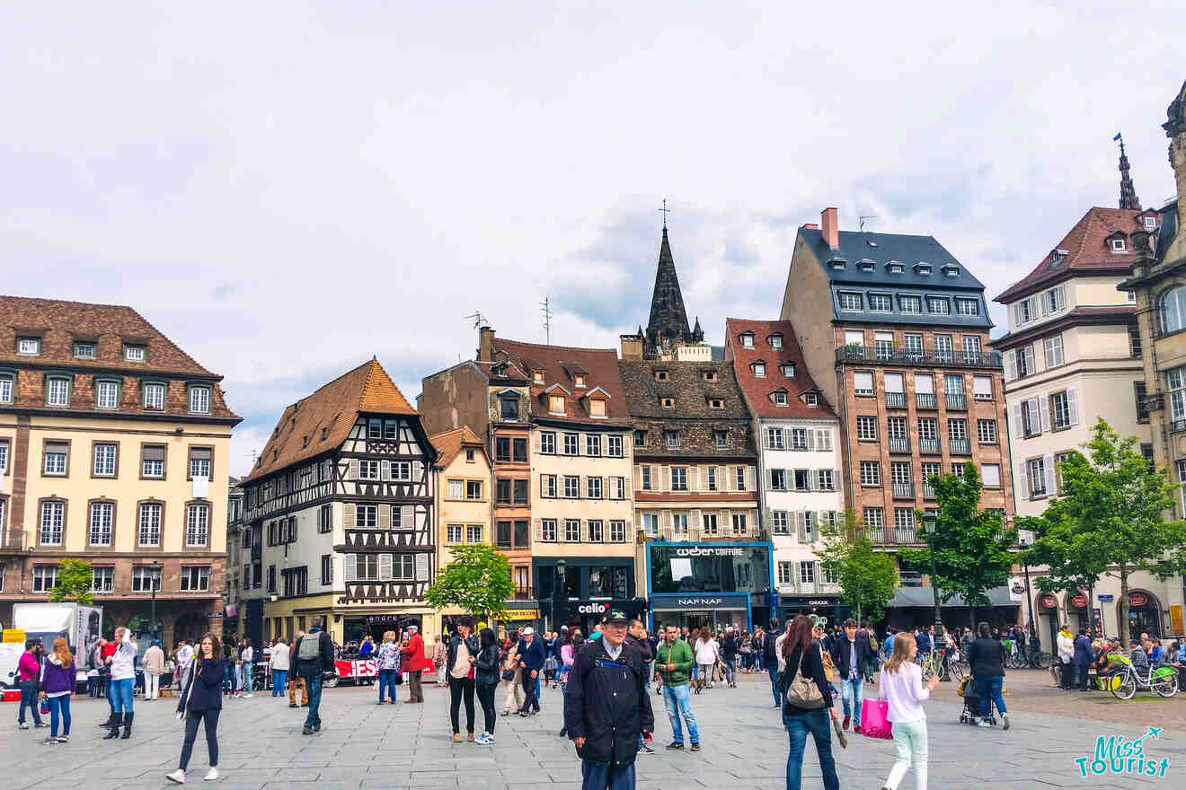 A bustling town square with people walking and historic buildings in the background under a cloudy sky.