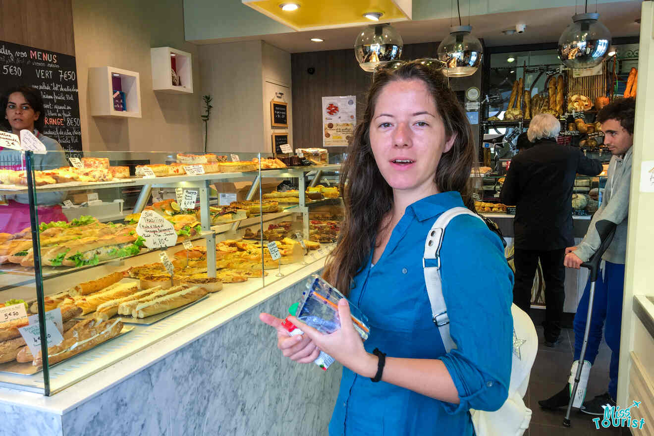 The writer of the post in blue shirt stands in front of a bakery display filled with pastries and sandwiches, holding a small map. Behind her, people browse the shop.