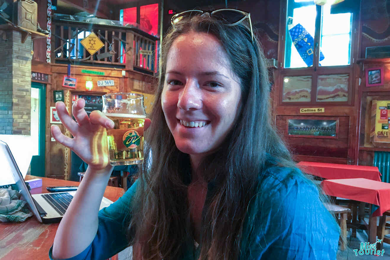 The writer of the post with long hair and glasses on their head holds a glass of beer in a pub setting, with colorful decor in a Parisian cafe