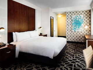 Hotel room with a large bed, wooden headboard, patterned wallpaper, abstract wall art, two side tables, and a desk with a chair.