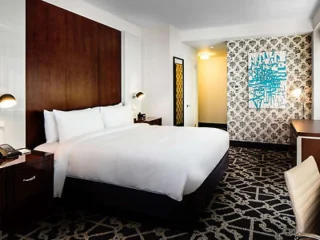 Hotel room with a large bed, wooden headboard, patterned wallpaper, abstract wall art, two side tables, and a desk with a chair.