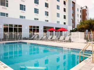 Outdoor hotel swimming pool with a white fence, surrounded by lounge chairs and red umbrellas. A multi-story building is in the background.