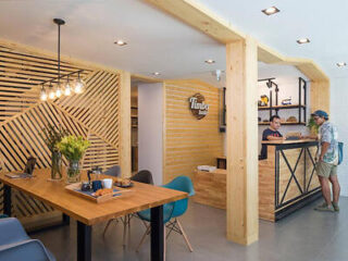 Modern cafe interior with wooden accents, a long table with chairs on the left, and a counter with two people on the right. Bright lighting and a decorative wall add to the ambiance.