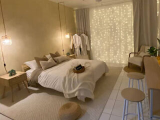 Cozy bedroom with a bed, decorative pillows, hanging lights, and a curtain of string lights. A small chair and stool are in the corner, and robes hang near the window.