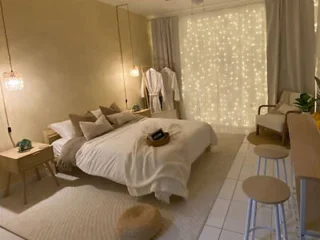 Cozy bedroom with a bed, decorative pillows, hanging lights, and a curtain of string lights. A small chair and stool are in the corner, and robes hang near the window.