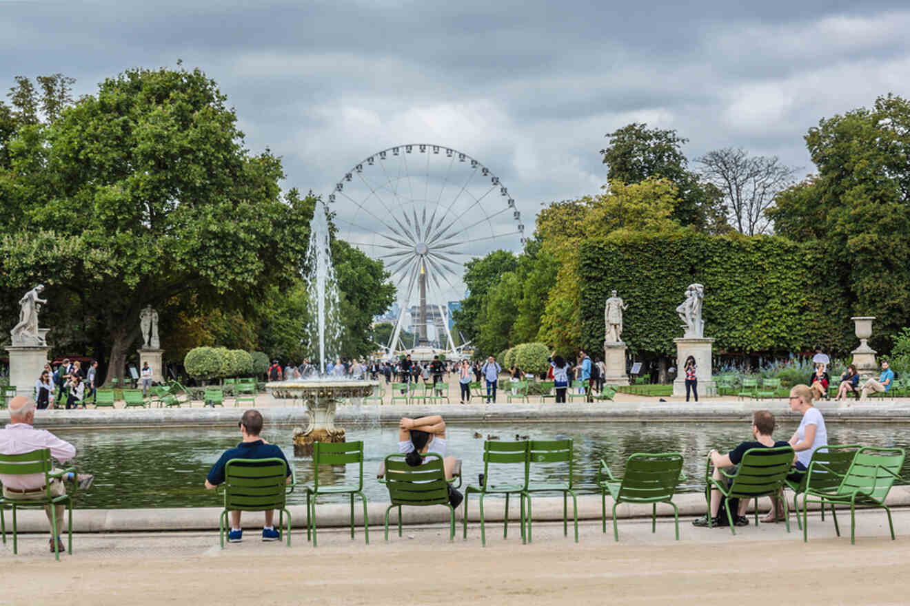 People sit on green chairs by a fountain in a park, with a ferris wheel and statues in the background under a cloudy sky.