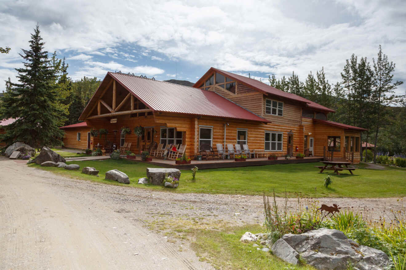 A large wooden cabin with a red roof and covered porch surrounded by grass, trees, and a gravel path under a partly cloudy sky.