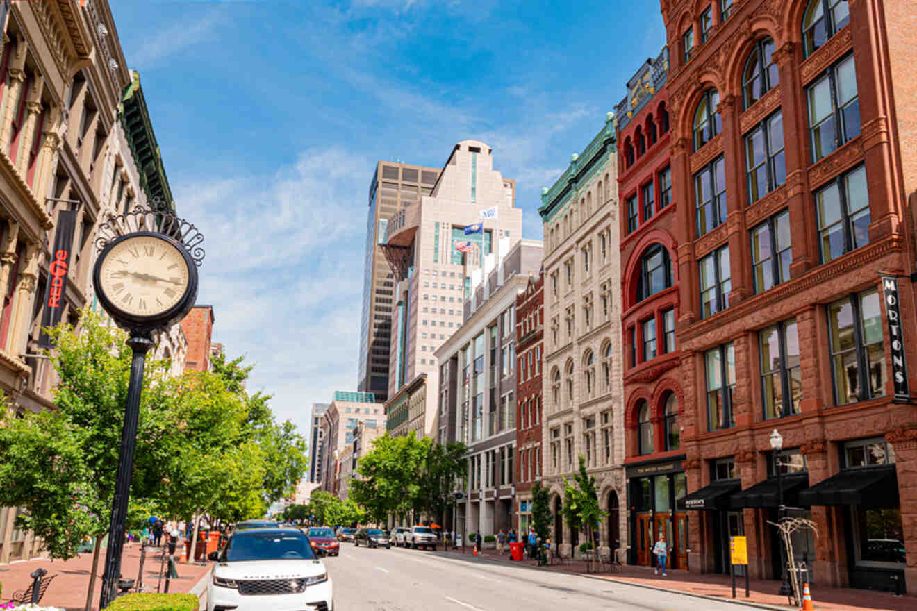 Street view of Louisville with historic buildings, a clock on a pole, trees lining the sidewalk, and a few cars parked on the road under a clear blue sky.