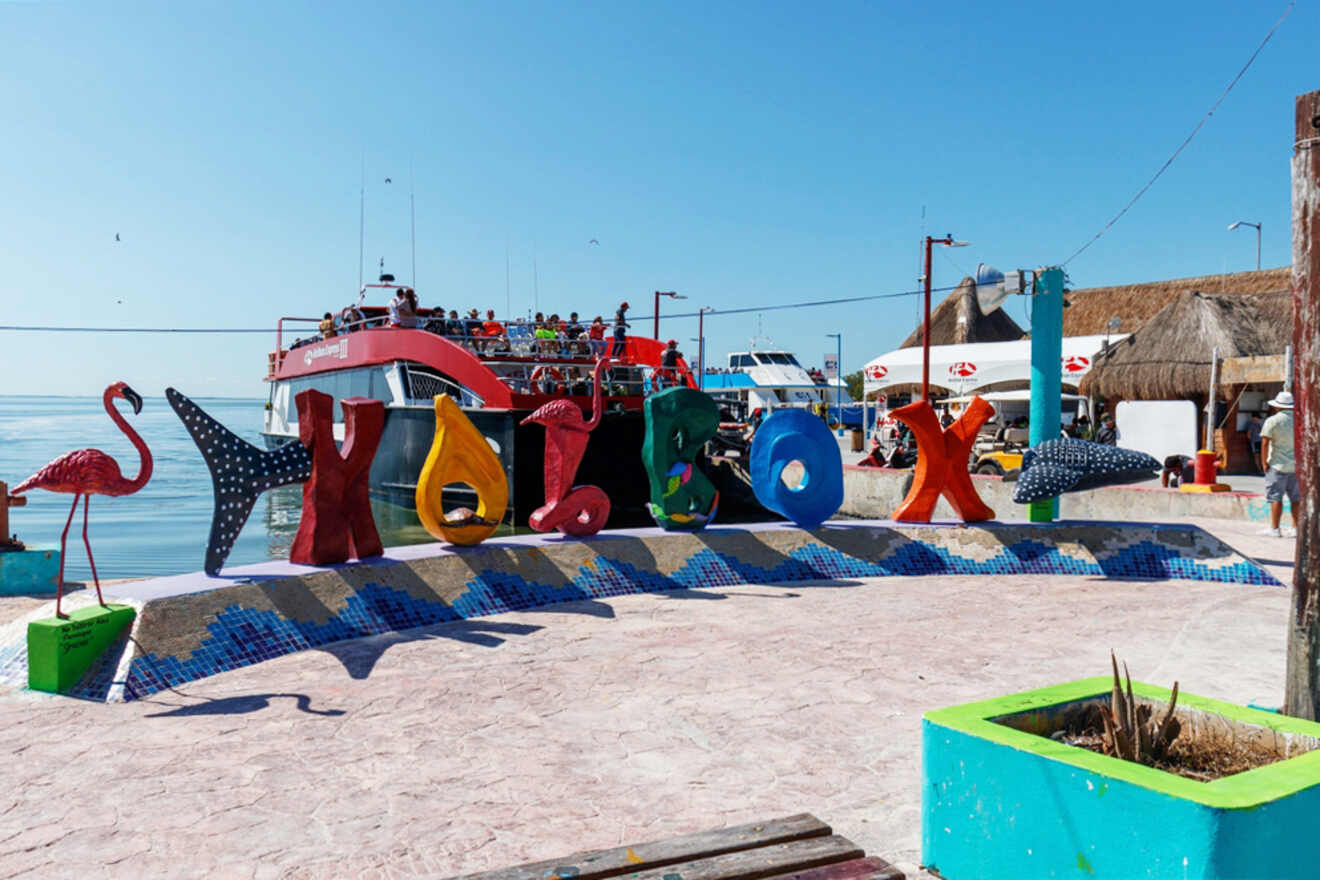 Colorful Holbox sign with decorative flamingo and whale shark figures, near a docked boat against a clear blue sky.