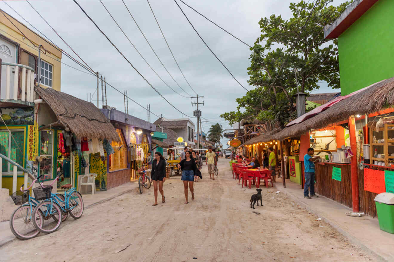 A bustling street scene with people walking, bicycles parked, and open-air restaurants with brightly colored facades. A dog roams the sandy road under an overcast sky.