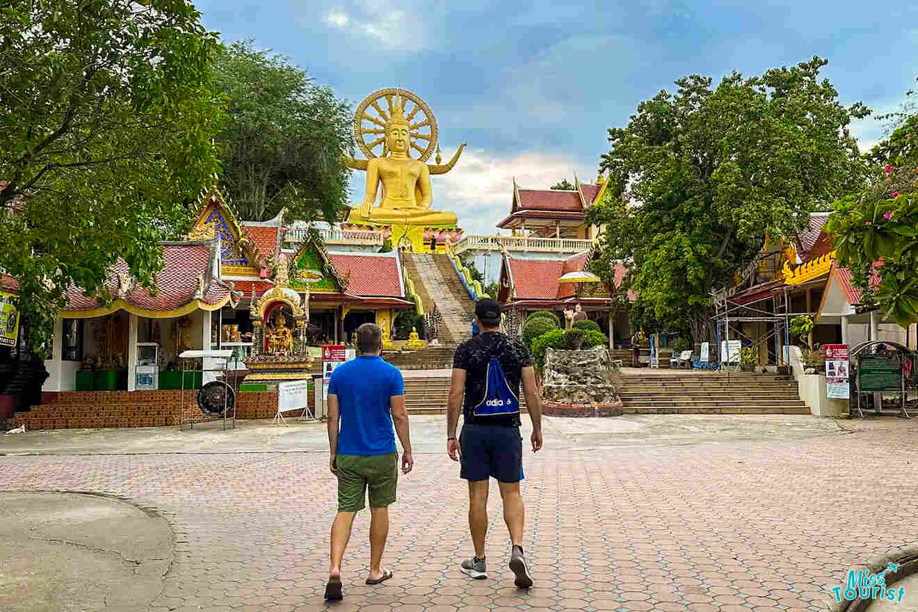 Two people walking toward a large golden Buddha statue in Thailand, surrounded by intricate temple architecture and trees.