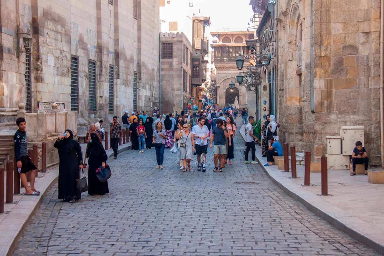 People walking on a cobblestone street in Old Cairo lined with historic buildings and street lamps.