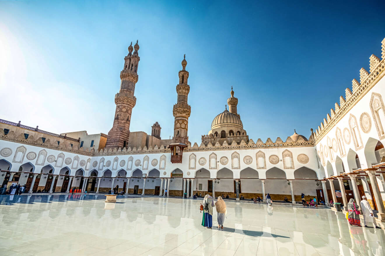 A bright day view of Al-Azhar Mosque's courtyard, featuring ornate minarets and a large dome. People walk across the shiny, open square surrounded by arcaded walls.