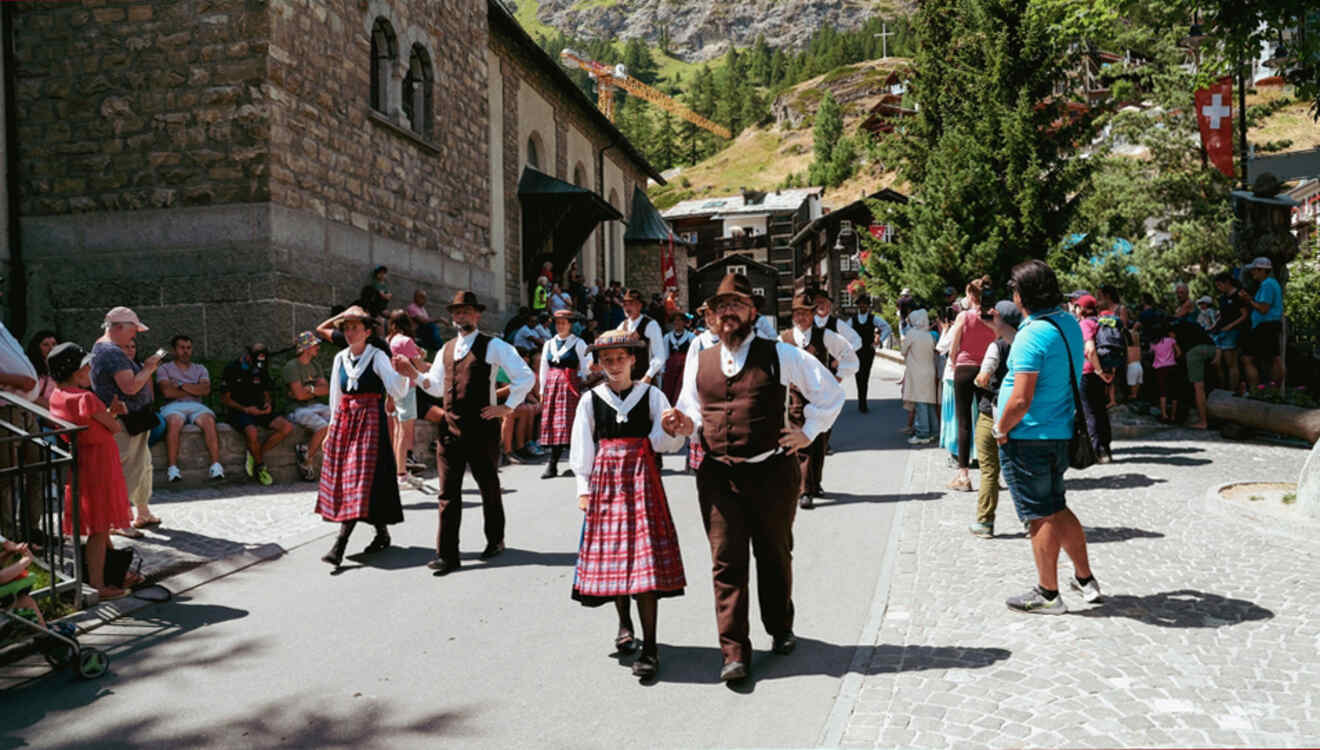 People in traditional clothing in Switzerland participate in a parade on a sunny day, with onlookers seated and standing along a cobblestone street.