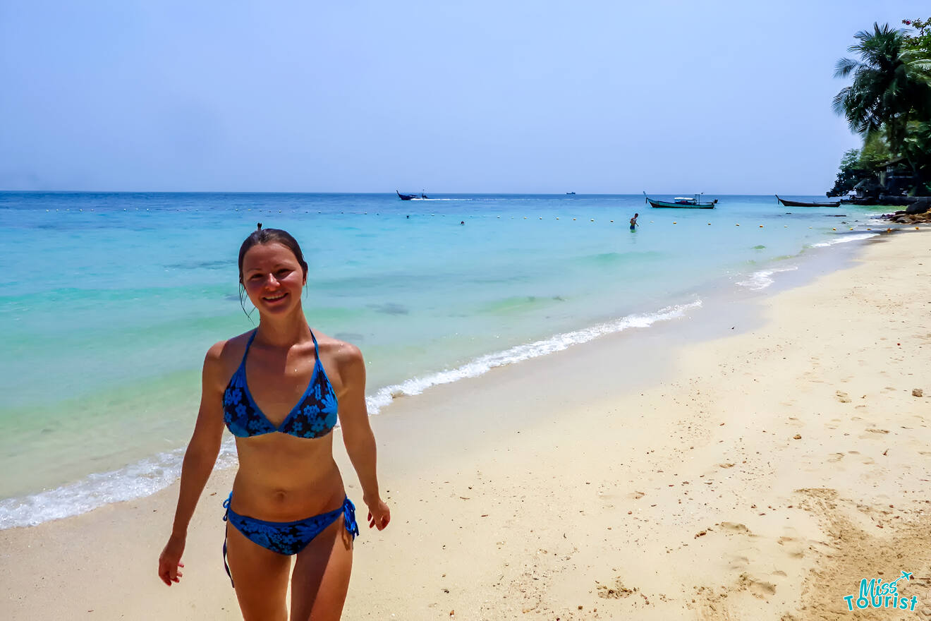 The writer of the post in a blue patterned swimsuit walks along a sandy beach with clear blue water and boats in the background inThailand