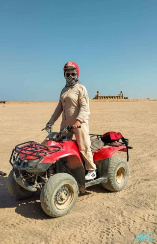 Person wearing protective gear riding a red ATV in a desert landscape. Sand and a building are visible in the background.