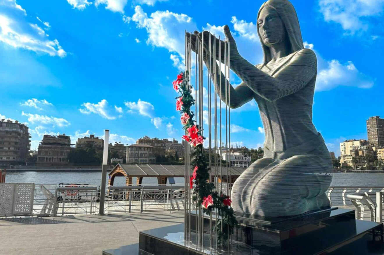 A large statue of a kneeling woman with harp-like strings, adorned with red flowers, set against a riverside backdrop with buildings and a bright blue sky.