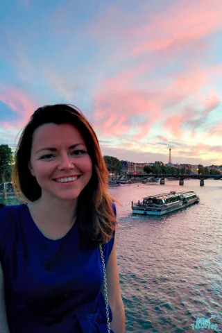 The writer of the post smiling in front of a river with a boat and a cityscape at sunset, featuring pinkish clouds.