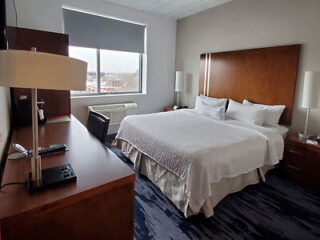 Hotel room with a large bed, two lamps, a desk, and a window with a city view. The room is simply decorated with neutral tones and wooden furniture.