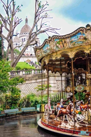 A vintage carousel sits in a park with the Sacré-Cœur Basilica visible in the background. Trees and benches surround the area on a cloudy day.