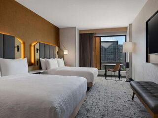 Hotel room with two double beds, a wall-mounted TV, a desk with a chair by the window, a bench, and a view of a building outside.