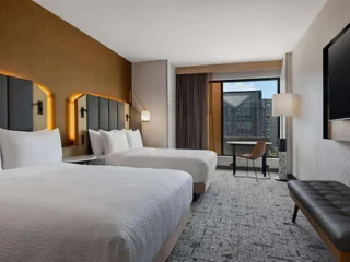 Hotel room with two double beds, a wall-mounted TV, a desk with a chair by the window, a bench, and a view of a building outside.