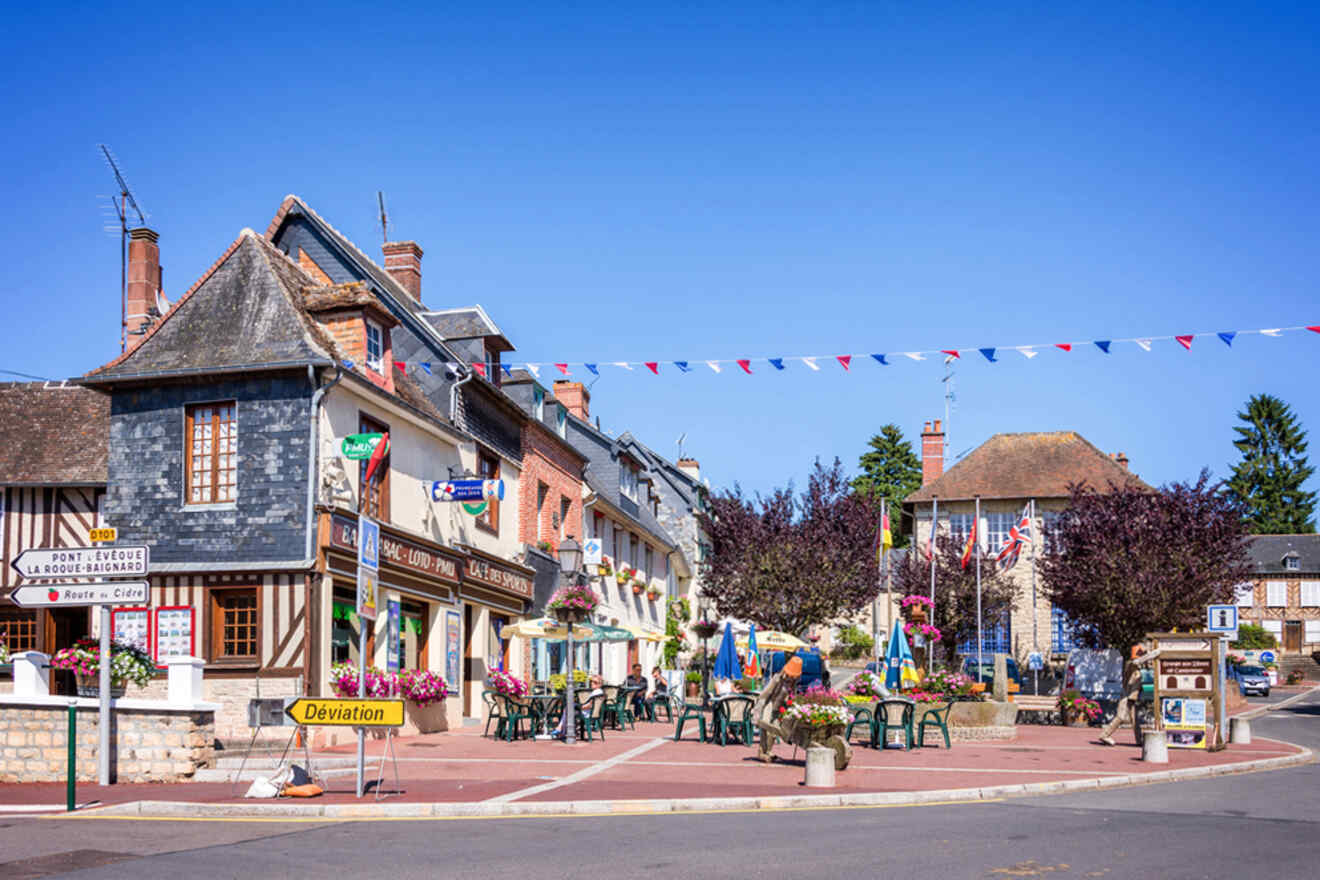 A quaint town square with traditional buildings, outdoor seating, and colorful flags. Bright blue sky above.