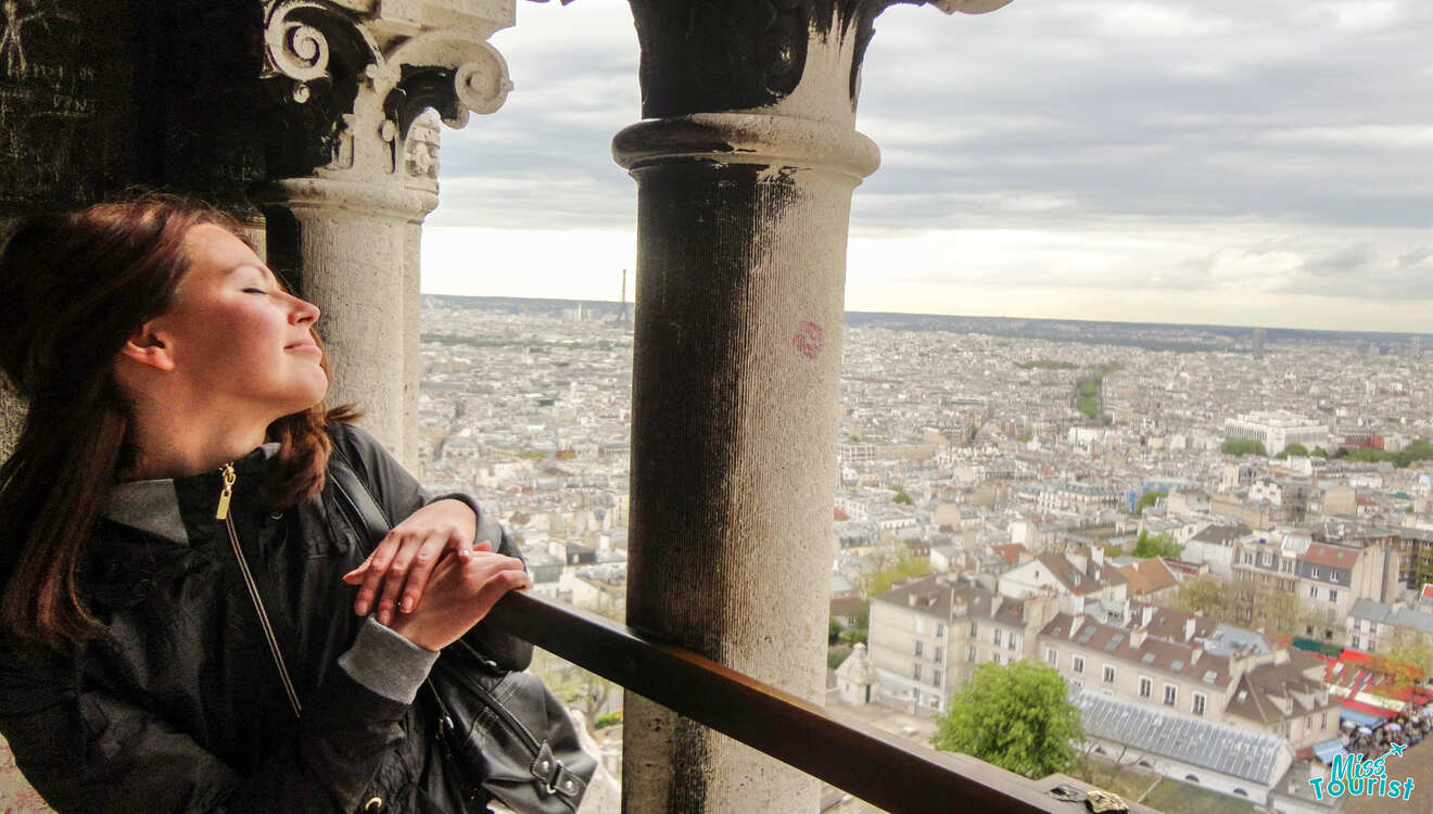 A woman relaxes against a stone railing, eyes closed, enjoying the view of a cityscape from a high vantage point on a cloudy day.