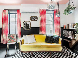 A living room with a yellow sofa, red patterned curtains, and a black-and-white rug. The walls have a two-tone design with art and plants.