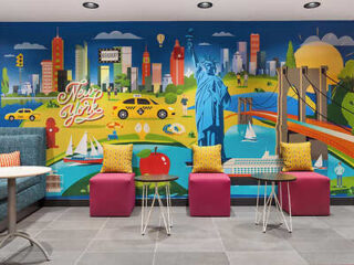 Colorful mural of New York City with landmarks like the Statue of Liberty and Brooklyn Bridge, in a seating area with pink chairs and small tables.