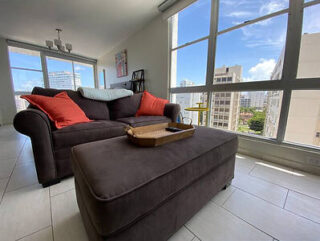 A cozy living room with a dark brown sofa, red and blue cushions, and a matching ottoman. Large windows reveal a cityscape with blue skies and high-rise buildings. A tray is on the ottoman.