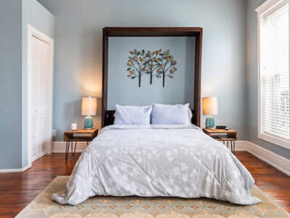 A neatly made bed with light bedding is between two wooden nightstands with lamps. A metal tree wall decoration is above the bed. The room has wooden floors and light blue walls.