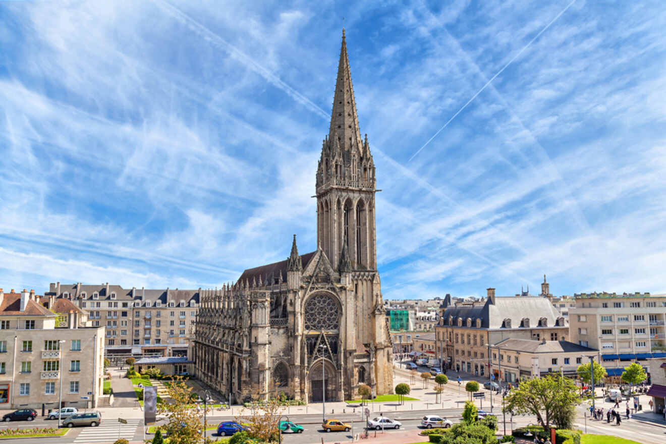 A Gothic-style cathedral with a tall spire stands in a town square surrounded by buildings and cars under a blue sky with streaks of clouds.