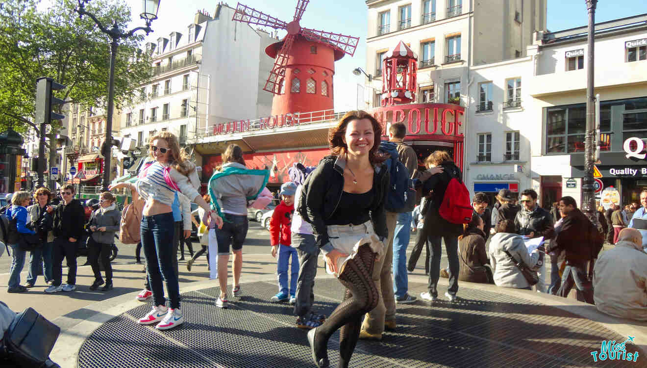 People posing near Moulin-Rouge and interacting with a windmill building in a busy urban area.