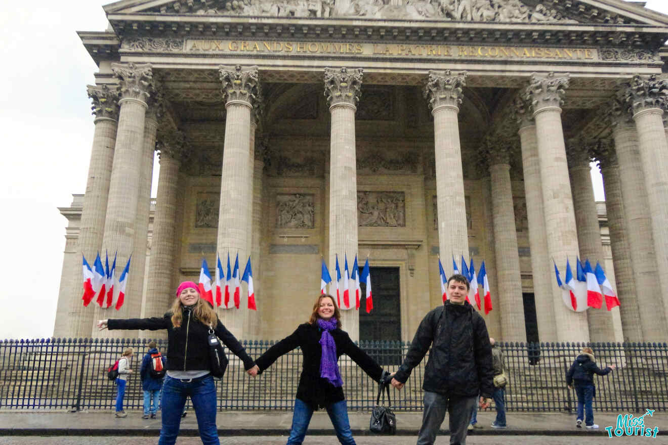 Three people holding hands in front of the Panthéon in Paris, decorated with French flags.