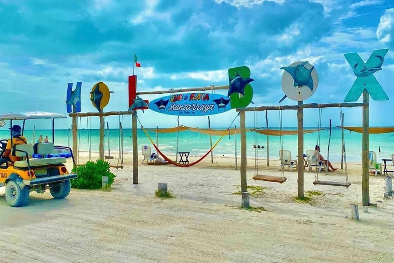 A beach scene with a sign reading "GXE Playa Santa Cruz." Swings and hammocks hang beneath the sign. A golf cart is on the sandy path. The ocean and blue sky are in the background.