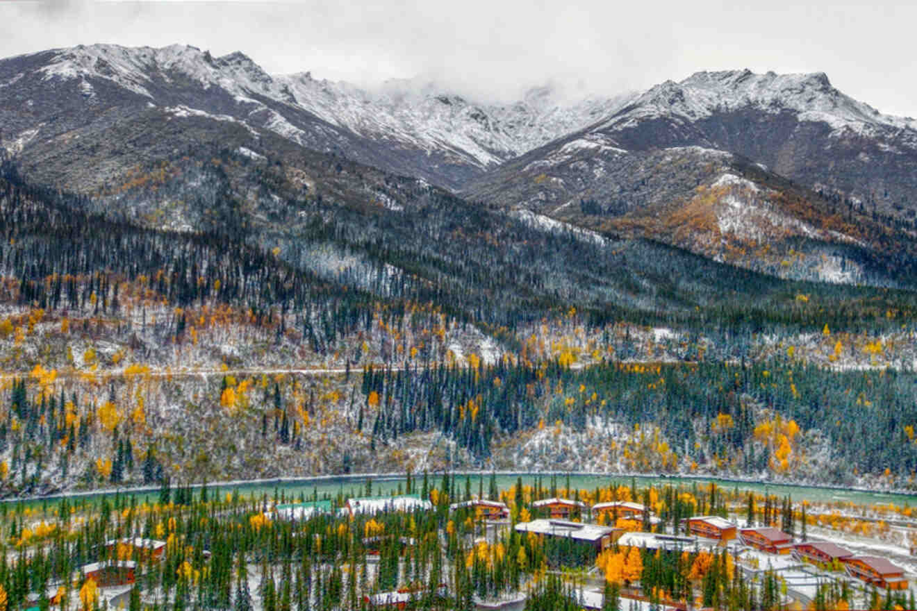 Colorful autumn forest with snow-dusted mountains and a few rustic buildings below.