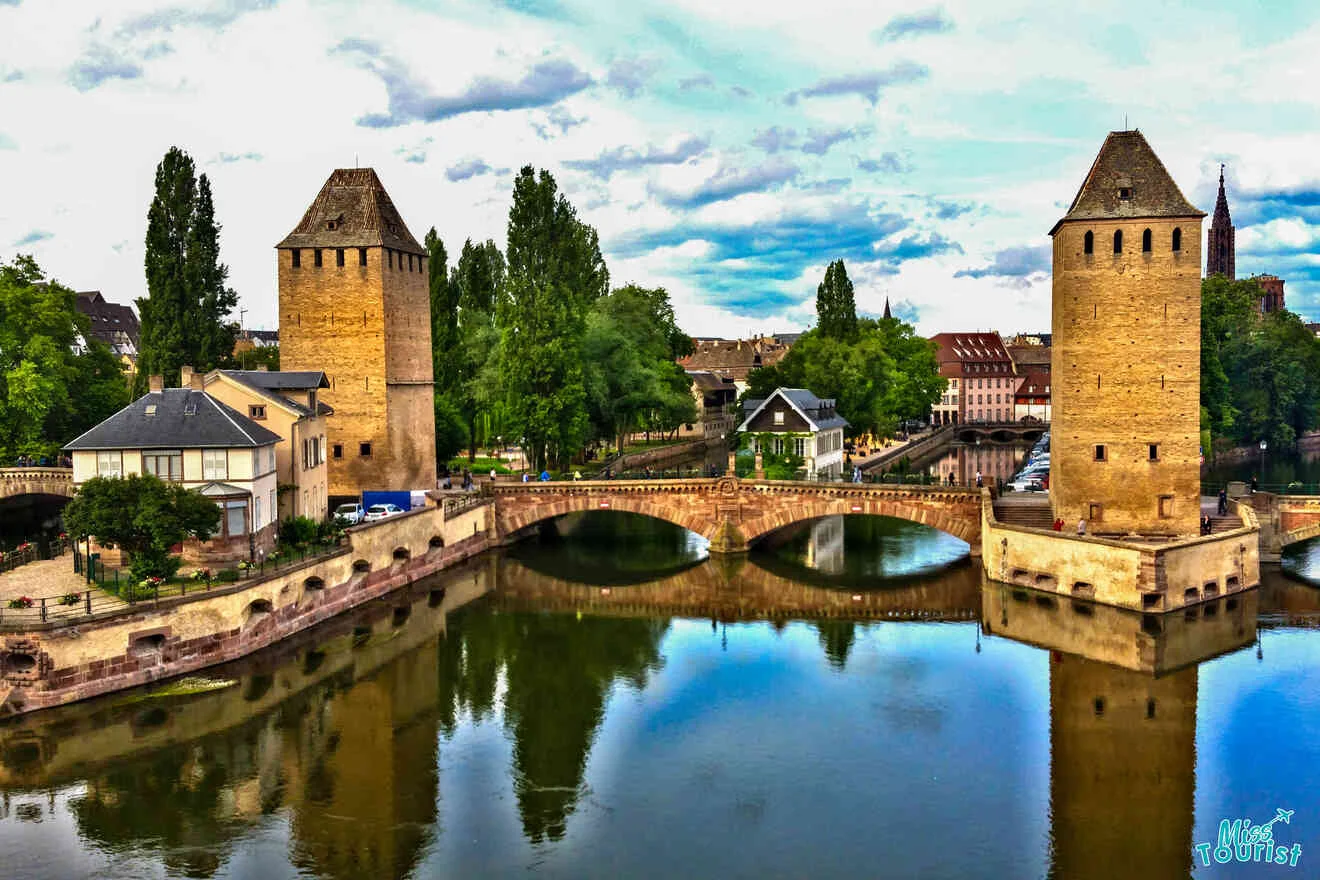 A scenic view of a historic stone bridge with three arches and two towers over a river. Trees and buildings are visible in the background under a partly cloudy sky.