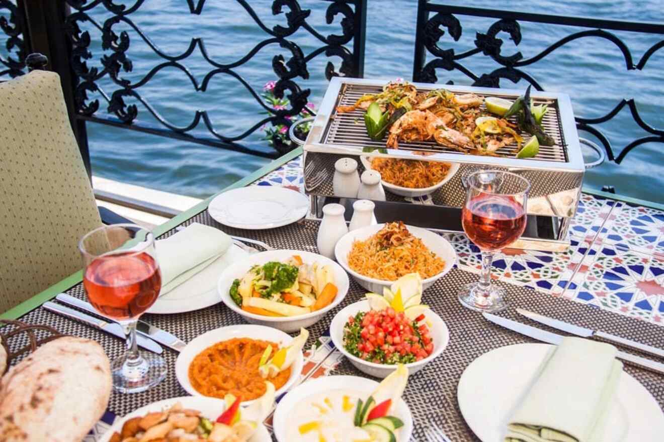 Outdoor dining setup with grilled seafood, various side dishes, and two glasses of rosé wine on a table by the water.