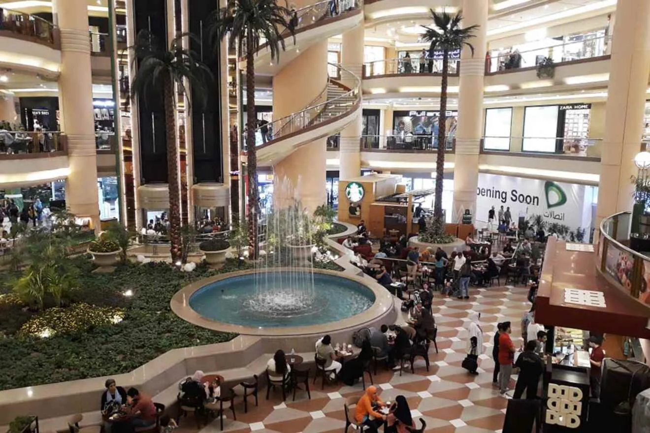 People sitting at tables around a central fountain in a multi-level shopping mall with palm trees and a sign that reads