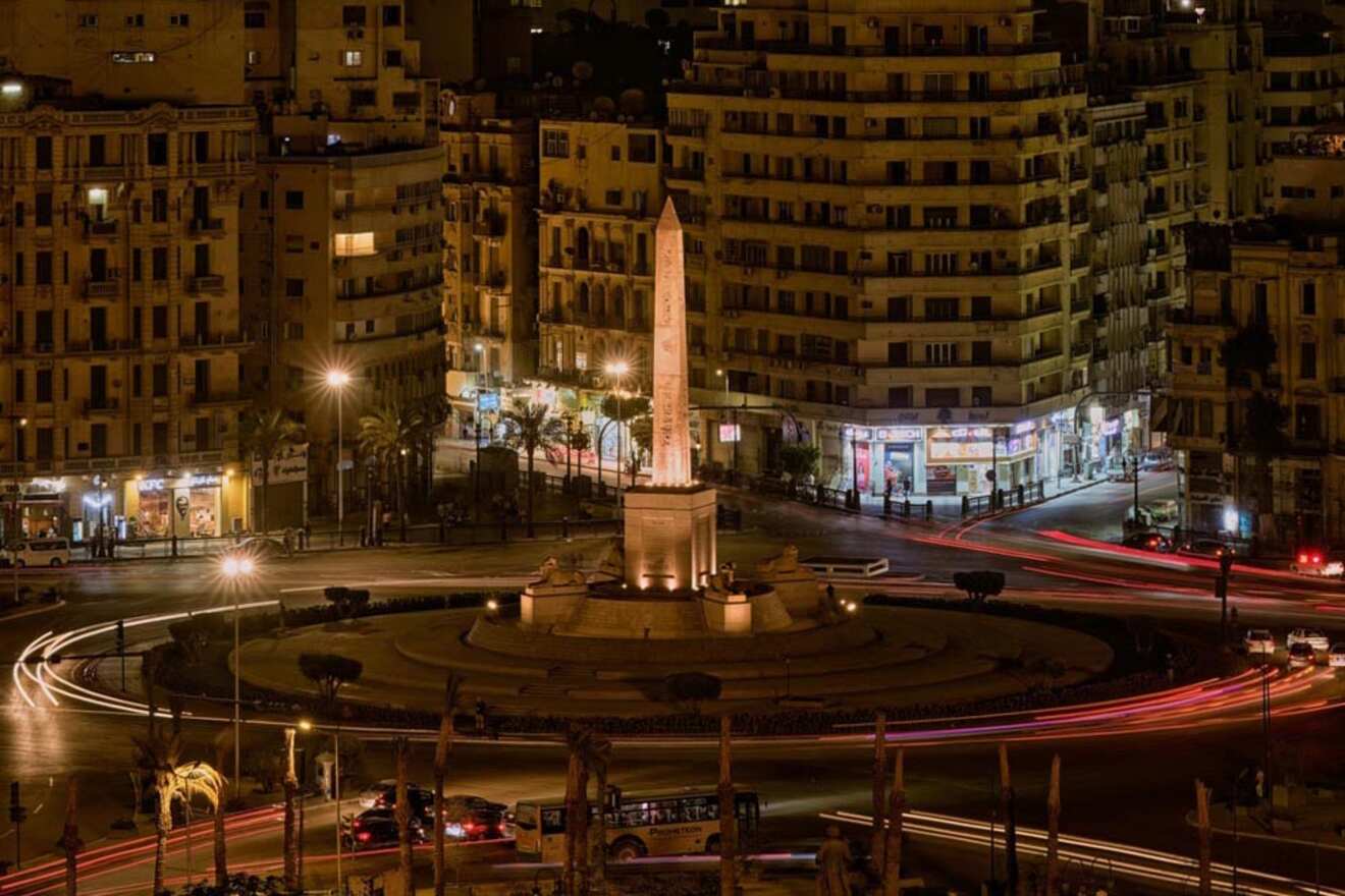 Night view of a lit obelisk in a circular roundabout surrounded by illuminated buildings and light trails from moving vehicles.