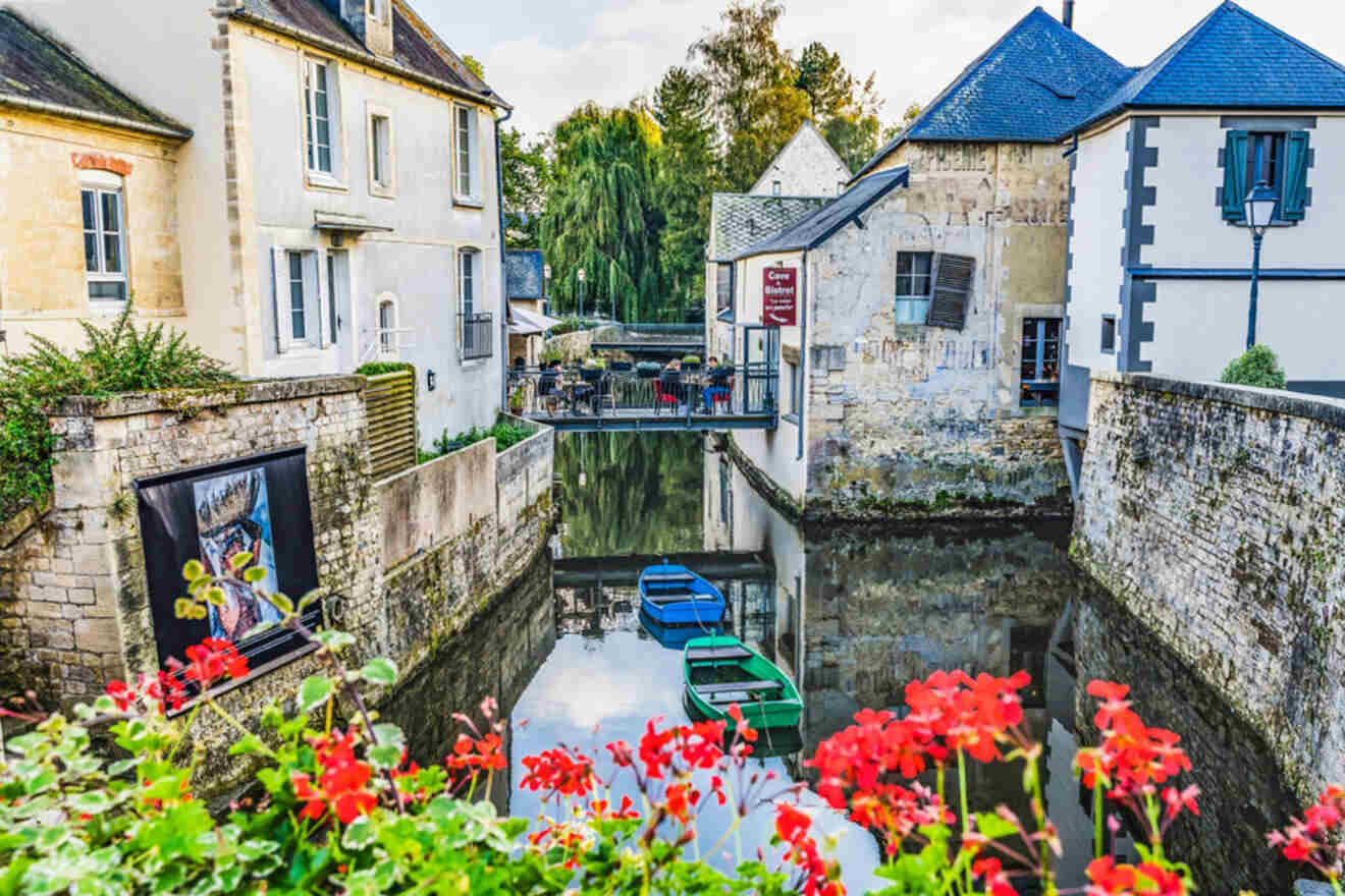 Quaint canal scene in Bayeux with small boats docked, surrounded by historic stone buildings and red flowers in the foreground.