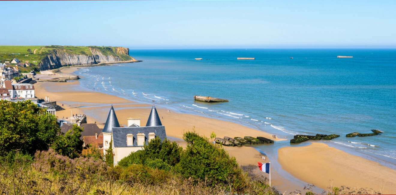 A coastal scene with sandy beach, cliffs, and scattered ruins in the water. A French flag is visible near beachfront buildings. Clear blue sky.